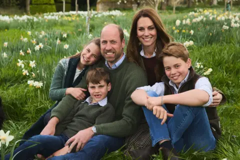 Kensington Palace The Prince and Princess of Wales with their three children sitting on the grass amongst some daffodils. Prince William is at the centre alongside Catherine. He is balding with short brwn hair and a cropped beard, and is wearing a green jumper with a light blue shirt collar visible. Prince Louis, who has a gapped toothed smile, is wearing similar clothes and is sitting in his lap. Leaning on his right shoulder is Princess Charlotte who is wearing a green jumper and dark tartan-style scarf. She has long brown hair and is siling at the camera. On his left shoulder is his wife, Princess Catherine, who is smiling. She has long brown hair and is wearing a deep red jumper. Her arm is round Prince George who is in blue jeans, a brown gilet and white shirt with his sleeves rolled up.