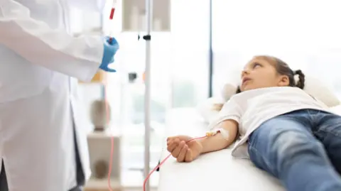 A small child having a blood transfusion. The child is wearing a white t-shirt and blue jeans. The child is laying down, and a medical professional is stood by them administering the blood transfusion.