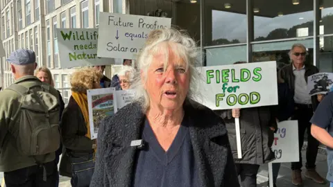 Rose Barnecut. She has long grey hair and is wearing a dark coloured jacket. She is stood in front of a row of protesters. 