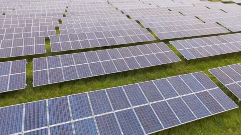 An aerial photo of a solar farm which is made up of rows of solar panels on top of a patch of grass.