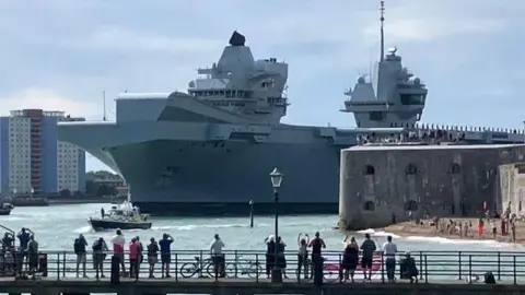 The large carrier close to the shore next to a tug boat with people gathered to watch it on its journey