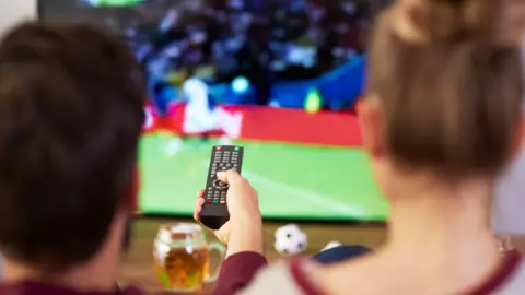 A man and woman seen from behind watching football on a television. The man is holding up a remote control and a glass of beer is on the table in front of him.