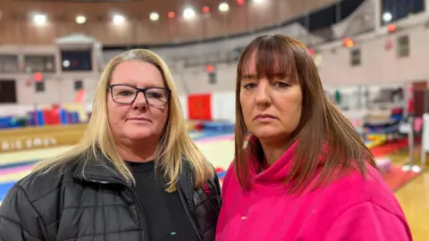 Julia Falle is standing to the left of her sister Mel Le Boutillier. They are both looking seriously at the camera. They are standing in front of an out-of-focus gym hall.