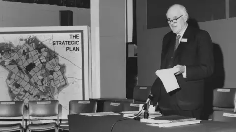 Getty Images Black-and-white photograph of John Campbell in a dark suit standing beside a table and microphone during a presentation. The person is holding papers and speaking in front of a large display board showing a map titled “THE STRATEGIC PLAN”. Rows of chairs with name labels are arranged behind the table, and documents are spread across the tabletop in a conference or meeting-room setting.