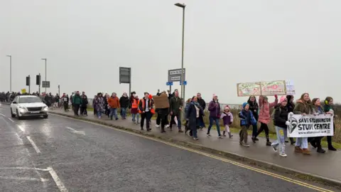 Protesters march along the pavement alongside a main road holding banners. They are made up of adults, children, and some dogs.