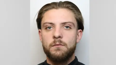 Wiltshire Police Mugshot of a man with brown slicked back hair and a beard. He looks into the camera with a serious expression.