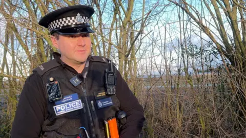 An image of PC Jonathan Lightfoot from Cheshire Police's rural crime team. He wears a black police uniform with equipment on it, a badge saying "Police" and stands in front of some trees.