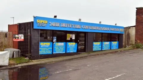 The front of the drive thru store, which is located inside two shipping containers which have been joined together. The shops sign reads 'Super Stop 24HR drive thru convenience store and vape shop'.