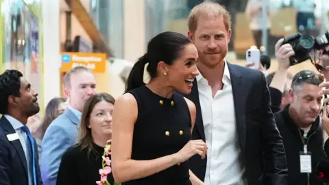 Harry and Meghan greet fans and press in Australia. Meghan is smiling and animated. She is wearing a navy top with gold buttons
