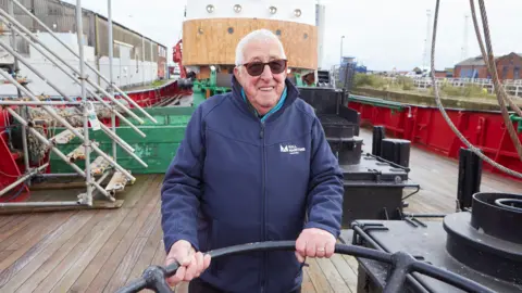Hull City Council An man with short white hair and sunglasses holds onto a black metal bar on the deck of a boat.