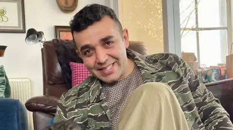 Family handout An Afghan man with short dark hair sitting in a leather chair in a living room to the side of a window, wearing a green camouflage-style jacket