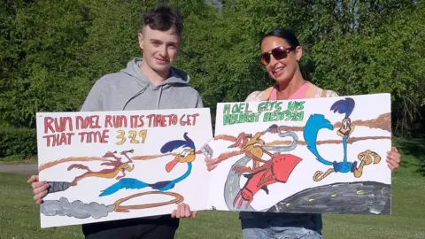 A man and women holding a white banner each. He is wearing a grey hoodie and black jeans and has dark hair. The lady has black hair with sunglasses with a multicoloured top and blue jeans.