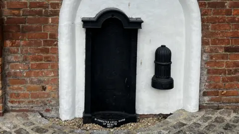 A close up of the fountain. It is a white painted arch on a red brick wall. Within the arch there is a cast iron back-plate with an inscription on the trough at street level, that reads Gloucester Local Board Of Health 1863. There is also a small black hydrant on the right. 