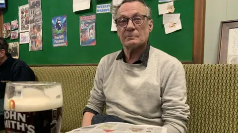BBC/Lucy Ashton A man with greying hair and glasses is wearing a black shirt with a grey sweatshirt over the top. He is sitting in a social club bar with a newspaper and a pint of beer on a table in front of him