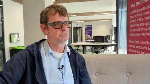 The image shows a Senior Lecturer in Economics at the University of Bournemouth. He is sat on a beige sofa. Behind him are two white desks with black office chairs. To the right of him is a pink banner. He has short brown hair and black glasses. He is wearing a blue shirt with a white top underneath. Over the top, he has a blue hoodie. 