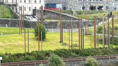 an array of steel pillars stand upright in an area of grass. a railway line is in the foreground