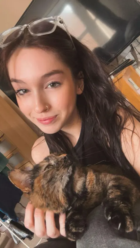 Rieve Nesbitt-Marr Ms Nesbitt-Marr in a black top with long, dark hair, smiles into the camera while stroking her tortoiseshell cat, Whiskey