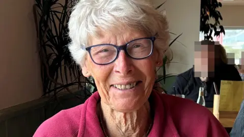 Jenny Firth is smiling as she she sits in a restaurant. She has quite short white hair and brown eyes and she is wearing a pair of glasses and a bright pink fleece. Behind her is a plant and a man sitting at another table whose face has been pixellated.
