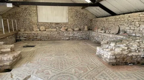 P Bates The mosaic room. In the centre is the mosaic floor which uses blue-grey, red, yellow and cream colours. The pattern has a knot-like design. The room has stone walls and a raised viewing platform.