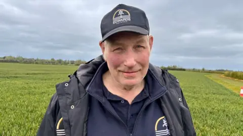 Dave Edwards is standing in a field of winter wheat in Lincolnshire. He is wearing a navy top, navy jacket and a baseball cap as he smiles at the camera.