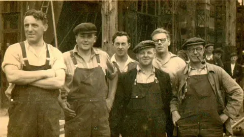 A black and white photograph of six workers at the Goole Shipyard. The men are wearing overalls and are smiling at the camera. Three of them are wearing flat caps and one is smoking a pipe.