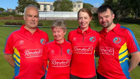 Four people, two men and two women dressed in red t-shirts standing on a bowling green.