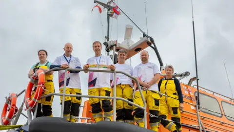 Vannin Photos Six members of a lifeboat crew are standing with yellow and black trousers and white t-shirts - they are looking down over the railings of a lifeboat on a cloudy day. The man third from the left is holding the baton.