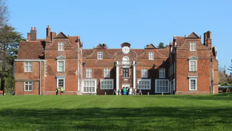 Ipswich Borough Council Christchurch Mansion is in the middle with a stretch of grass in front of it. The building is made of red bricks, and has multiple windows that are painted white. A group of people are standing outside the entrance which is in the middle.