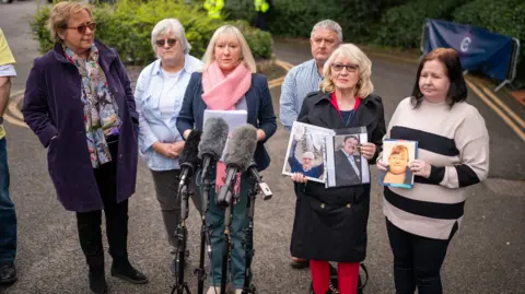 PA Media Five women and a man stand in front of microphones, with two women holding large photographs showing people's faces.