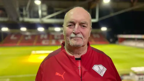 Michael Bull smiles at the camera in front of Lincoln City's football pitch He is wearing a red football tracksuit and is smiling at the camera. Mr Bull has a bald head and a grey goatee beard. 