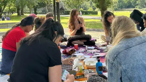 A group of women sitting in a park on a picnic rug. They have lots of cards and bowls scattered on the rug as they paint inside cards