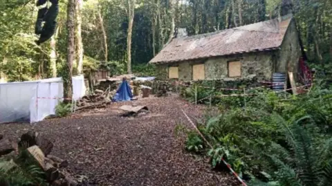 Io ACC Planning Documents The near derelict-looking cottage with boarded windows in a small clearing surrounded by trees