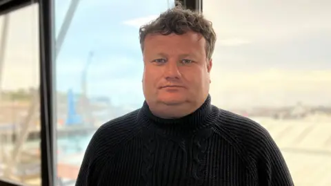 William is standing indoors in front of large windows, wearing a dark knit sweater. He has short brown hair and is looking directly at the camera.