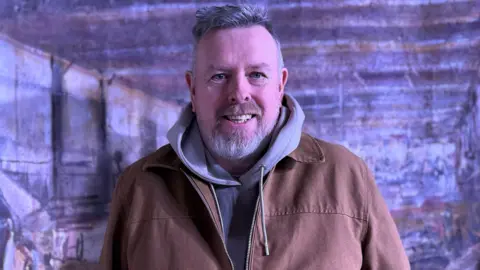 Sam is smiling. He's wearing a grey hoodie under a brown jacket. He's standing in front of a backdrop showing a factory scene.