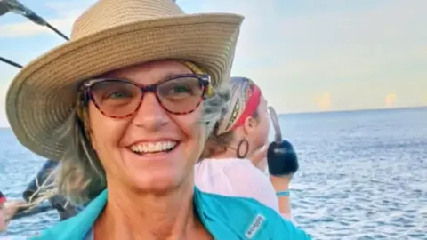 A photo of Lynette Hooker smiling while on a body of water. She is wearing a turquoise-like coloured shirt, glasses, and a woven straw hat.