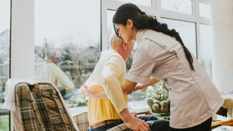 A care home worker helps an elderly resident down into a chair
