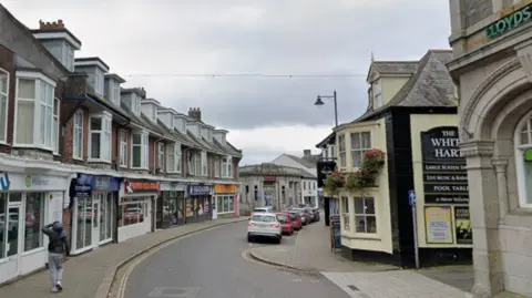 A general view looking down a street in a town centre. Shops line each side of the street. Cars are parked in on street car parking bays. A person is walking along a pavement.