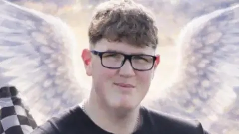 An image of a young man with glasses and short curly brown hair. He is looking at the camera and smiling. Behind him are some animated angel wings, a black and white chequered racing flag and a tyre.