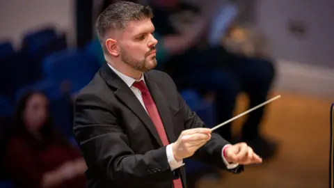 Christopher Bond A man called Christopher Bond conducts a band (who are out of vision). He is wearing a black suit with a white shirt and red tie. He has black trimmed hair and a black trimmed beard.