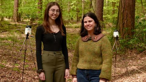 Two women in their twenties stand in front of two air sampling machines surrounded by woodland.