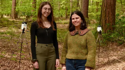 BBC/Martin Giles Two women in their twenties stand in front of two air sampling machines surrounded by woodland.