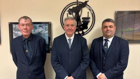 Three smartly dressed men in navy blue suits and ties are standing in front of the crest of Comhairle Nan Eilean Siar. It is a birlinn style medieval ship with a sail and oars.