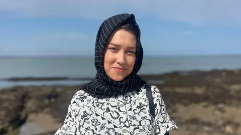 BBC Omulbanin Sultani is smiling straight at the camera. She is only visible from the chest up. She is wearing a black headscarf covering her hair with thin white pinstripes on it. She is wearing a white top with black pattern on it and has the strap of a black handbag over her left shoulder. She is standing in front of a blurred coastal background.