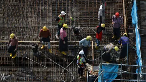 Getty Images Migrant Workers, usando capacetes amarelos e camisas de mangas compridas, fiquem em um canteiro de obras em 3 de maio de 2023 em Cingapura