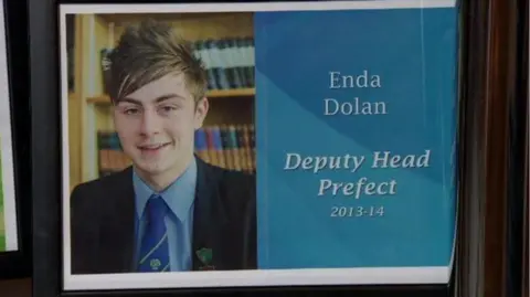 Dolan family A school photo of the late Enda Dolan. He is smiling and wearing a dark school blazer, blue shirt and school tie. The screen is split down the middle and on the other side there is a white graphic on a blue background which reads: "Enda Dolan, Deputy Head Prefect 2013-2014". 