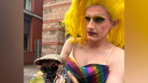 A person in drag with a bright yellow wig and full make up and rainbow dress holding a tiny dog in a flat cap and sunglasses