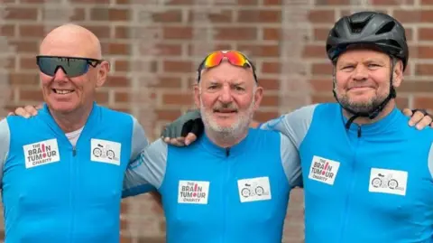 Steve Huntley/BBC The three dads are standing next to each other with their arms over each other's shoulders. They are all wearing a light blue T-shirt that says The Brain Tumour Charity on the right.