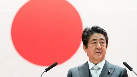 Getty Images De Japanse premier Shinzo Abe spreekt tijdens de diploma-uitreiking van de National Defense Academy op 22 maart 2020. De vlag van Japan is op de achtergrond