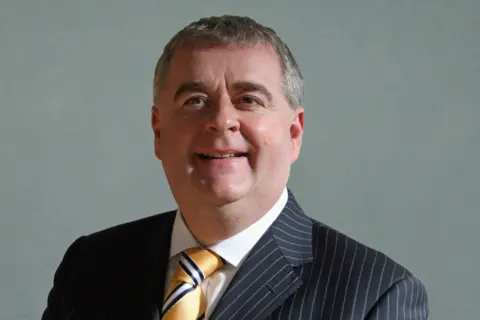 SBC A man with dark hair, wearing a dark jacket, white shirt and yellow tie looks at the camera