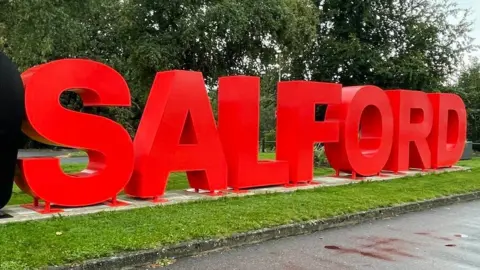 A large sign with red letters reading Salford stands on a grey concrete patch of ground, surrounded by green grass